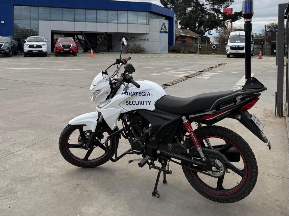 Moto de patrullaje blanca de Estrategia Security con baliza, estacionada frente a la fachada de una instalación